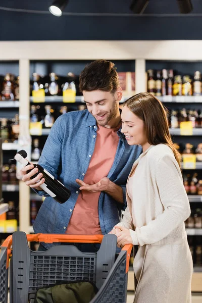 Coppia allegra guardando bottiglia di vino vicino al carrello della spesa nel supermercato — Foto stock