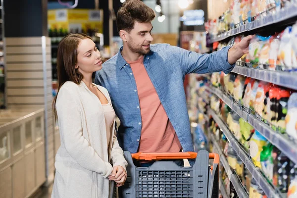 Jeune couple debout près du panier et des étagères avec épicerie au premier plan flou dans un supermarché — Photo de stock
