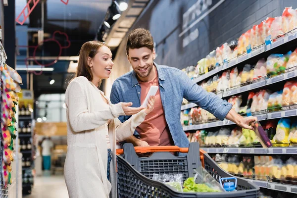 Mujer sonriente apuntando al teléfono inteligente cerca de novio y carrito de compras en el supermercado - foto de stock