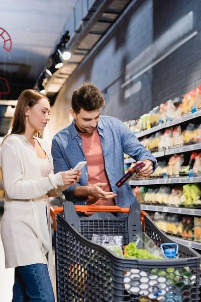 Woman holding smartphone near boyfriend with grocery and shopping cart on blurred foreground — Stock Photo