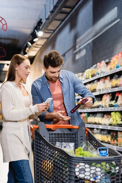 Donna che tiene smartphone vicino al fidanzato con generi alimentari e carrello della spesa in primo piano sfocato — Foto stock