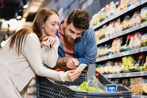 Smiling woman holding smartphone near boyfriend in supermarket — Stock Photo