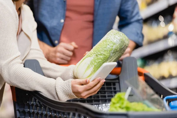 Обрізаний вид жінки, що тримає салат і смартфон біля хлопця на розмитому фоні в супермаркеті — стокове фото