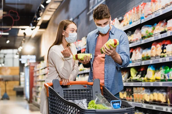 Donna in maschera medica che punta con mano vicino fidanzato con pacchetti di generi alimentari in negozio — Stock Photo