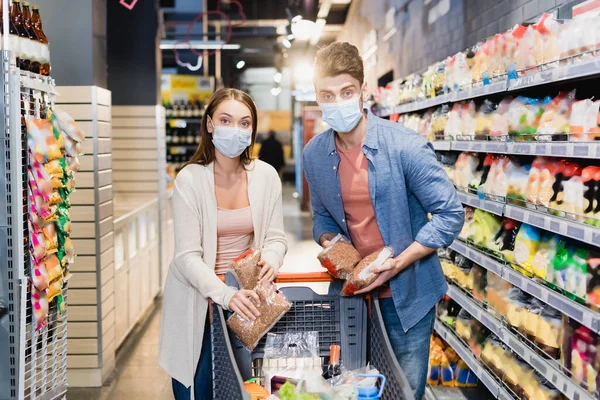 Pareja en máscaras médicas sosteniendo paquetes con trigo sarraceno en el supermercado - foto de stock