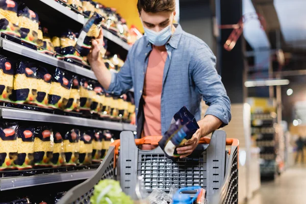 Uomo in maschera medica prendere la spesa vicino scaffali e carrello della spesa nel supermercato — Stock Photo