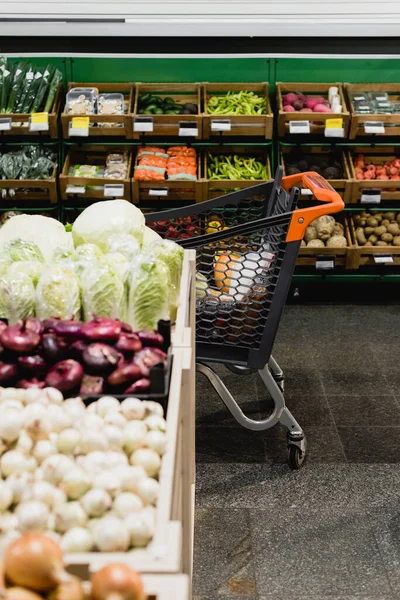 Shopping cart near fresh vegetables on blurred foreground in supermarket — Stock Photo