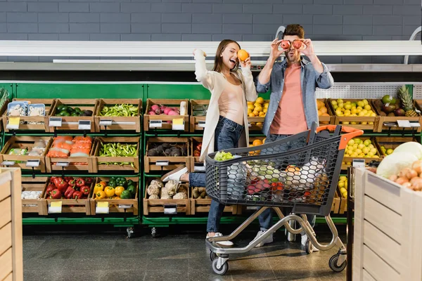 Fröhliches Paar hält Früchte in der Nähe von Gesichtern und Einkaufswagen im Supermarkt — Stockfoto