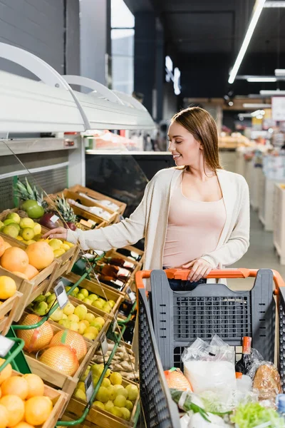 Donna allegra che prende pompelmo vicino a frutta e carrello della spesa nel supermercato — Foto stock