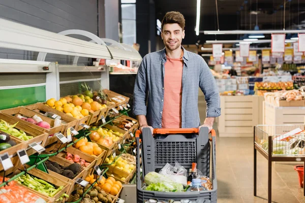 Hombre sonriente mirando la cámara cerca del carrito de la compra en el supermercado - foto de stock