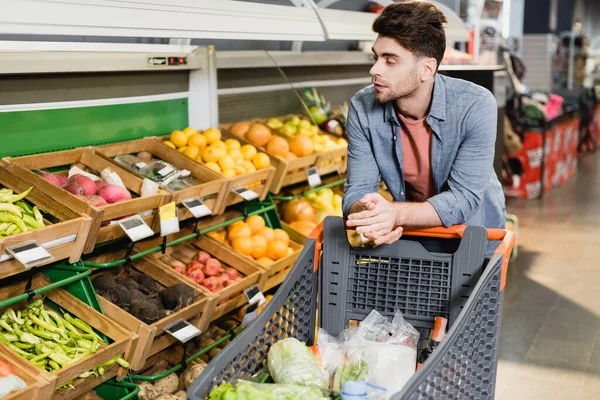 Young man standing near shopping cart and fresh fruits on counter in store — Stock Photo