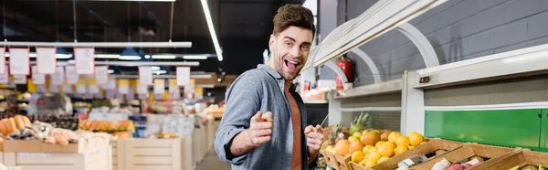 Fröhlicher Mann zeigt mit Fingern auf Obst im Supermarkt, Transparent — Stockfoto