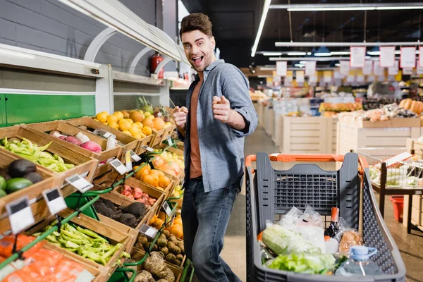 Homme joyeux pointant avec les doigts à la caméra près du chariot d'achat et de la nourriture dans le supermarché — Photo de stock