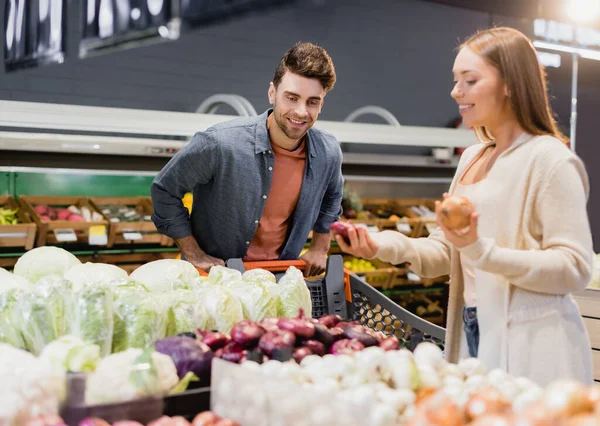 Uomo sorridente in piedi vicino al carrello della spesa e fidanzata con cipolle in primo piano sfocato nel supermercato — Foto stock