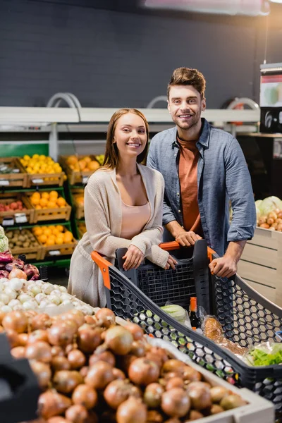 Casal sorridente em pé perto de carrinho de compras e legumes no supermercado — Fotografia de Stock