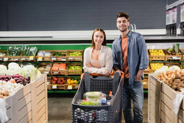 Young couple standing near shopping trolley and vegetables in supermarket — Stock Photo