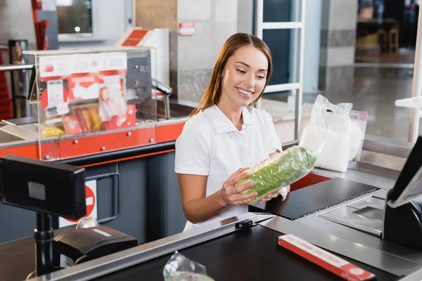 Smiling cashier holding lettuce on supermarket checkout — Stock Photo