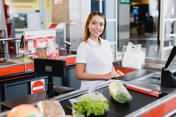 Lächelnde Kassiererin blickt in die Kamera neben den Lebensmitteln an der Supermarktkasse — Stockfoto