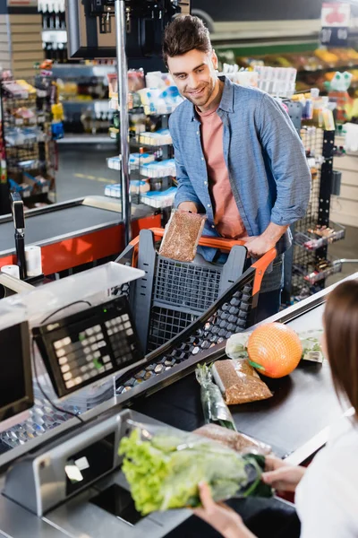 Sourire homme tenant paquet avec sarrasin près de la caisse sur la caisse du supermarché — Photo de stock