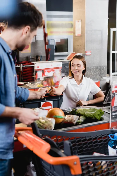 Cajero sonriente que toma la tarjeta de crédito del cliente cerca de la comida en la caja del supermercado - foto de stock