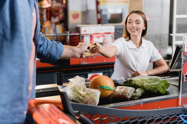 Homem dando cartão de crédito para caixa sorrindo perto de comida no checkout do supermercado — Fotografia de Stock