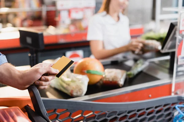 Vista recortada del hombre que sostiene la tarjeta de crédito cerca de la cesta de la compra y la compra del supermercado en el fondo borroso - foto de stock