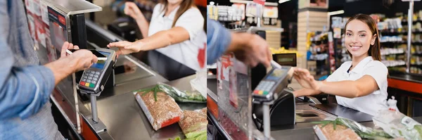 Collage de l'homme payant avec smartphone près de caissier souriant et de la nourriture sur la caisse du supermarché, bannière — Photo de stock