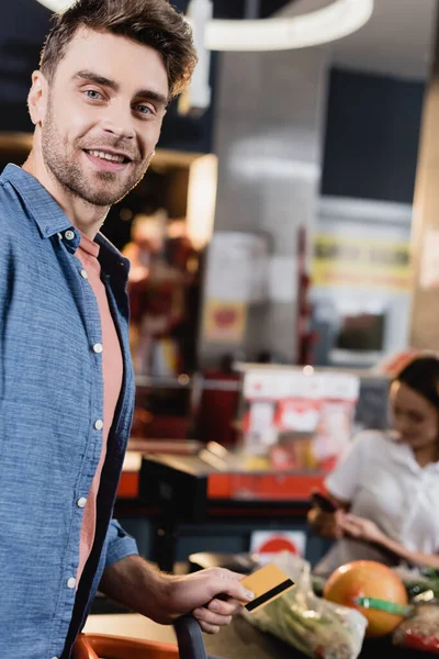 Cliente sonriente sosteniendo tarjeta de crédito cerca de la caja del supermercado en un fondo borroso - foto de stock