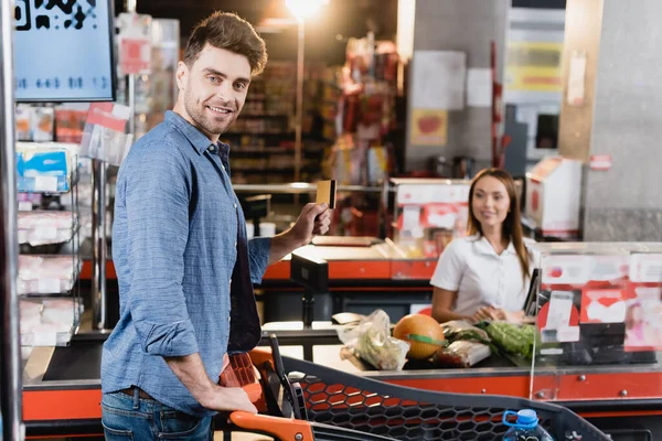 Sonriente hombre con tarjeta de crédito cerca del carrito de la compra y el supermercado de pago en un fondo borroso - foto de stock