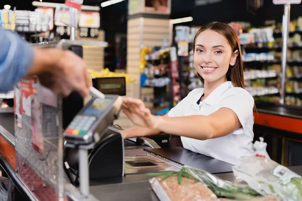 Smiling cashier looking at camera near customer paying with smartphone on blurred foreground in supermarket — Stock Photo