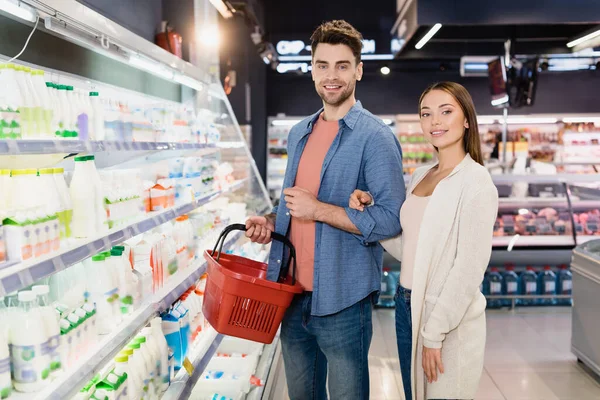 Smiling couple with shopping basket standing near food in supermarket — Stock Photo