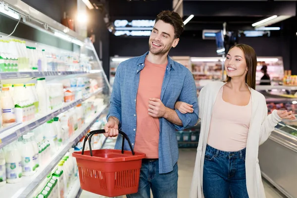 Pareja sonriente con cesta de la compra mirando la comida en primer plano borrosa en el supermercado - foto de stock