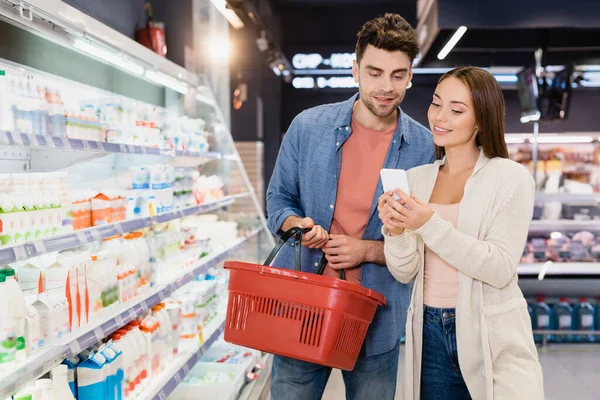 Woman using smartphone near boyfriend with shopping basket in supermarket — Stock Photo