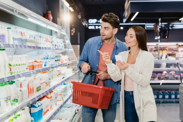 Woman with smartphone pointing with finger at food near boyfriend with shopping basket in supermarket — Stock Photo