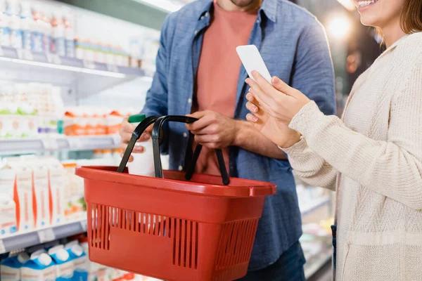 Ausgeschnittene Ansicht von Frau mit Smartphone in der Nähe ihres Freundes, der Flasche in Einkaufskorb legt — Stockfoto