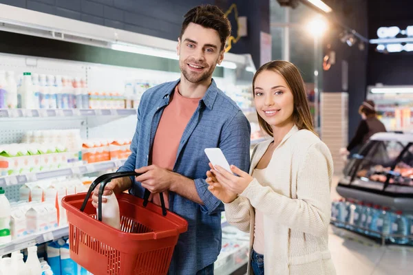 Pareja sonriente con smartphone y cesta de la compra mirando a la cámara en el supermercado - foto de stock