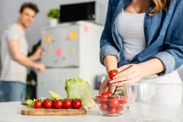 Vista recortada de la mujer sosteniendo tomate rojo cereza cerca de los ingredientes en la mesa de la cocina y novio sobre fondo borroso - foto de stock