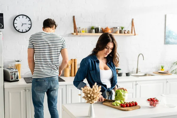 Frau schneidet Salat neben Zutaten auf Küchentisch und Freund — Stockfoto