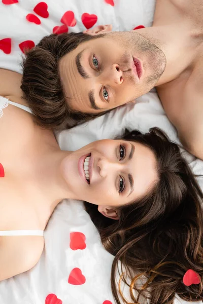 Vista superior do casal deitado na cama perto de corações de papel vermelho no dia dos namorados — Fotografia de Stock