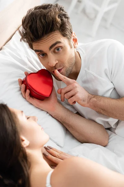 Man holding red heart-shaped box and showing hush sign near sleeping woman on blurred foreground — Stock Photo