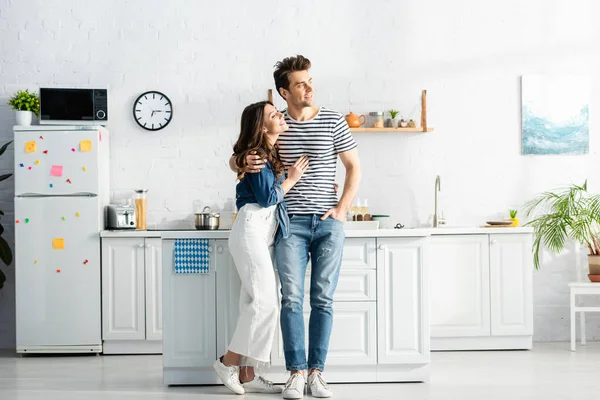 Comprimento total da mulher feliz abraçando homem alegre de pé com a mão no bolso na cozinha moderna — Fotografia de Stock