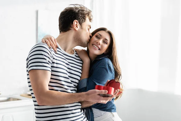 Homem beijando bochecha de feliz namorada enquanto segurando presente em forma de coração — Fotografia de Stock