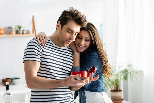 Hombre sorprendido mirando en forma de corazón caja de regalo cerca de novia feliz - foto de stock