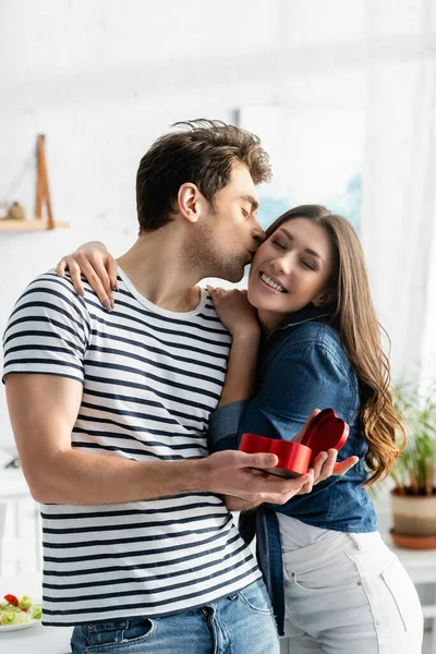 Homme embrassant joue de petite amie heureuse tout en tenant boîte cadeau en forme de coeur — Photo de stock