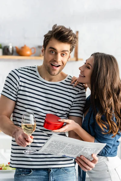 Homme étonné tenant un verre de vin et de journal près de petite amie joyeuse avec boîte cadeau en forme de coeur — Photo de stock