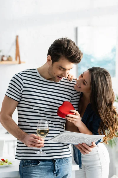 Happy man holding glass of wine and newspaper near cheerful girlfriend with heart-shaped gift box — Stock Photo