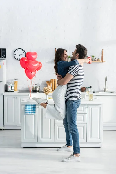 Full length of couple embracing near heart-shaped balloons in modern kitchen — Stock Photo