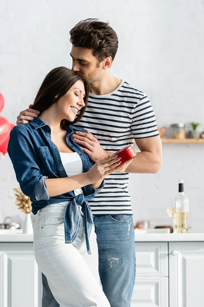 Man kissing head of happy girlfriend holding heart-shaped gift box — Stock Photo