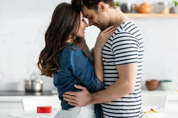 Vista lateral de pareja feliz con los ojos cerrados abrazándose en la cocina - foto de stock