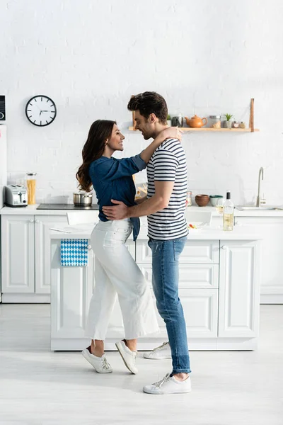 Piena lunghezza di uomo felice abbracciando fidanzata gioiosa e sorridente in cucina — Foto stock